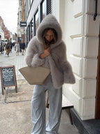 Woman in a grey hooded faux fur jacket and light blue pants holding a handbag