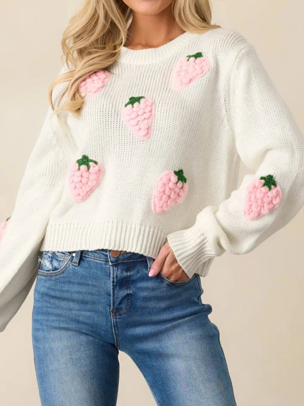 Front view of a woman in a white cropped knit sweater featuring all-over pink strawberry embroidery, paired with blue denim jeans.