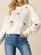 Front view of a woman in a white cropped knit sweater featuring all-over pink strawberry embroidery, paired with blue denim jeans.