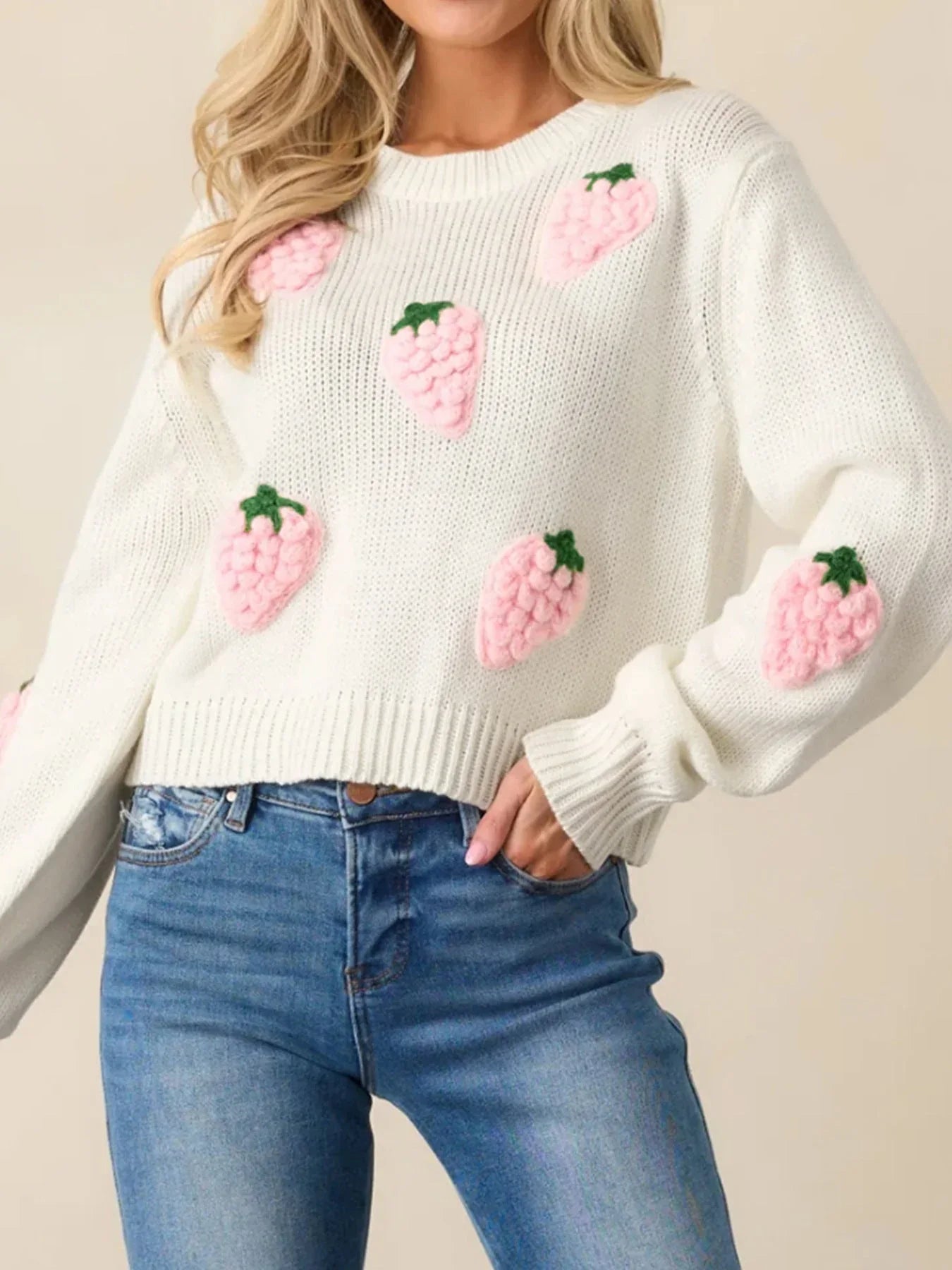 Front view of a woman in a white cropped knit sweater featuring all-over pink strawberry embroidery, paired with blue denim jeans.