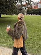 Woman from behind wearing a green faux fur jacket and carrying a tan crossbody bag while walking in a park.