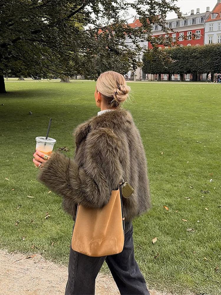 Woman from behind wearing a green faux fur jacket and carrying a tan crossbody bag while walking in a park.