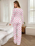 Rear view of a woman wearing a white long-sleeve pajama top and matching pants with a pink heart print, standing in a bedroom.