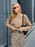 Back view of a woman wearing a tan textured knit sweater with long sleeves and a curved hem, paired with blue jeans.