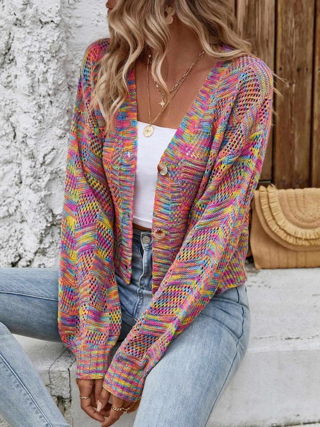 Woman sitting down wearing a rainbow multicolor open-knit cropped cardigan with a v-neck and button-front closure, styled with a white top and blue jeans.