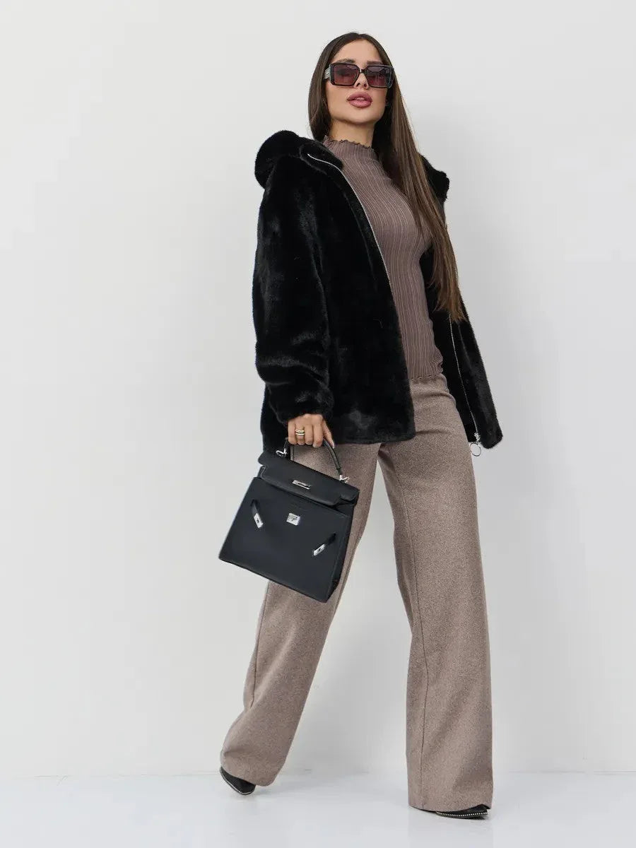 Full body shot of a woman wearing a black hooded faux fur jacket, taupe ribbed knit top, and taupe wide-leg pants, accessorized with sunglasses and a black handbag.