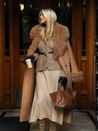 Woman in a long camel coat with shearling trim, holding a coffee cup and a brown handbag.