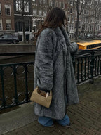 Woman wearing a long grey faux fur coat with lapel collar and full length sleeves, standing by a canal.