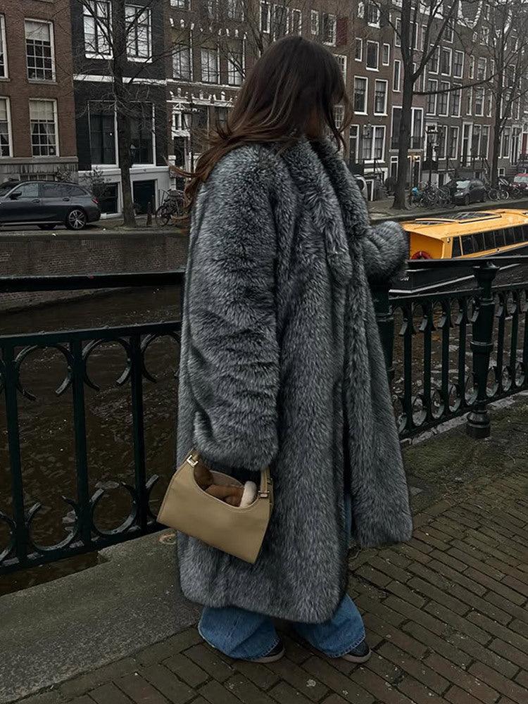 Woman wearing a long grey faux fur coat with lapel collar and full length sleeves, standing by a canal.