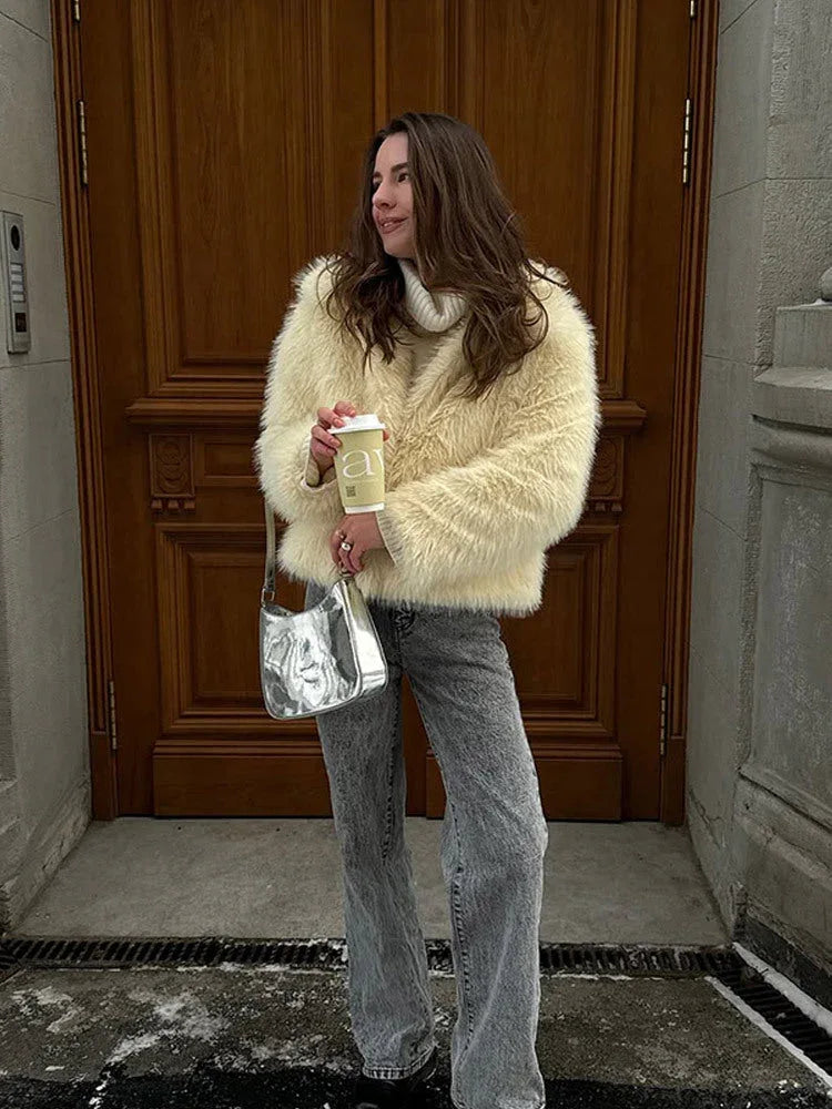 Woman with a silver handbag, wearing a cream faux fur jacket over a white turtleneck sweater and grey jeans, standing in front of a wooden door.