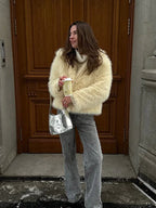 Woman with a silver handbag, wearing a cream faux fur jacket over a white turtleneck sweater and grey jeans, standing in front of a wooden door.