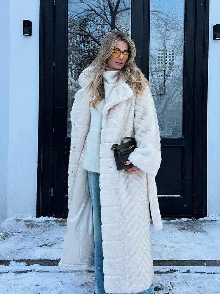 Woman wearing a long, cream-colored faux fur coat with a chevron pattern and a belted waist, standing outdoors in the snow.