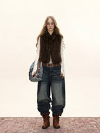 Woman wearing a brown faux fur vest, white long-sleeve shirt, wide-leg denim pants, a studded belt, and brown boots, with a shoulder bag.