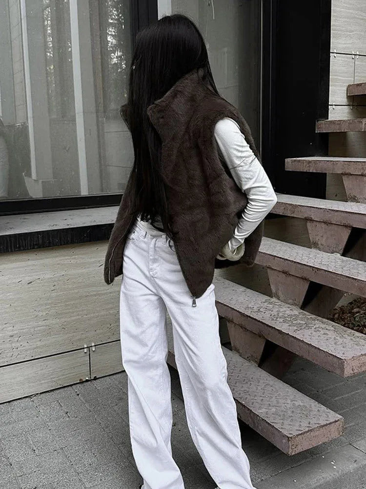 Woman wearing a brown faux fur vest over a white long-sleeved shirt and white wide-leg jeans, standing outdoors.