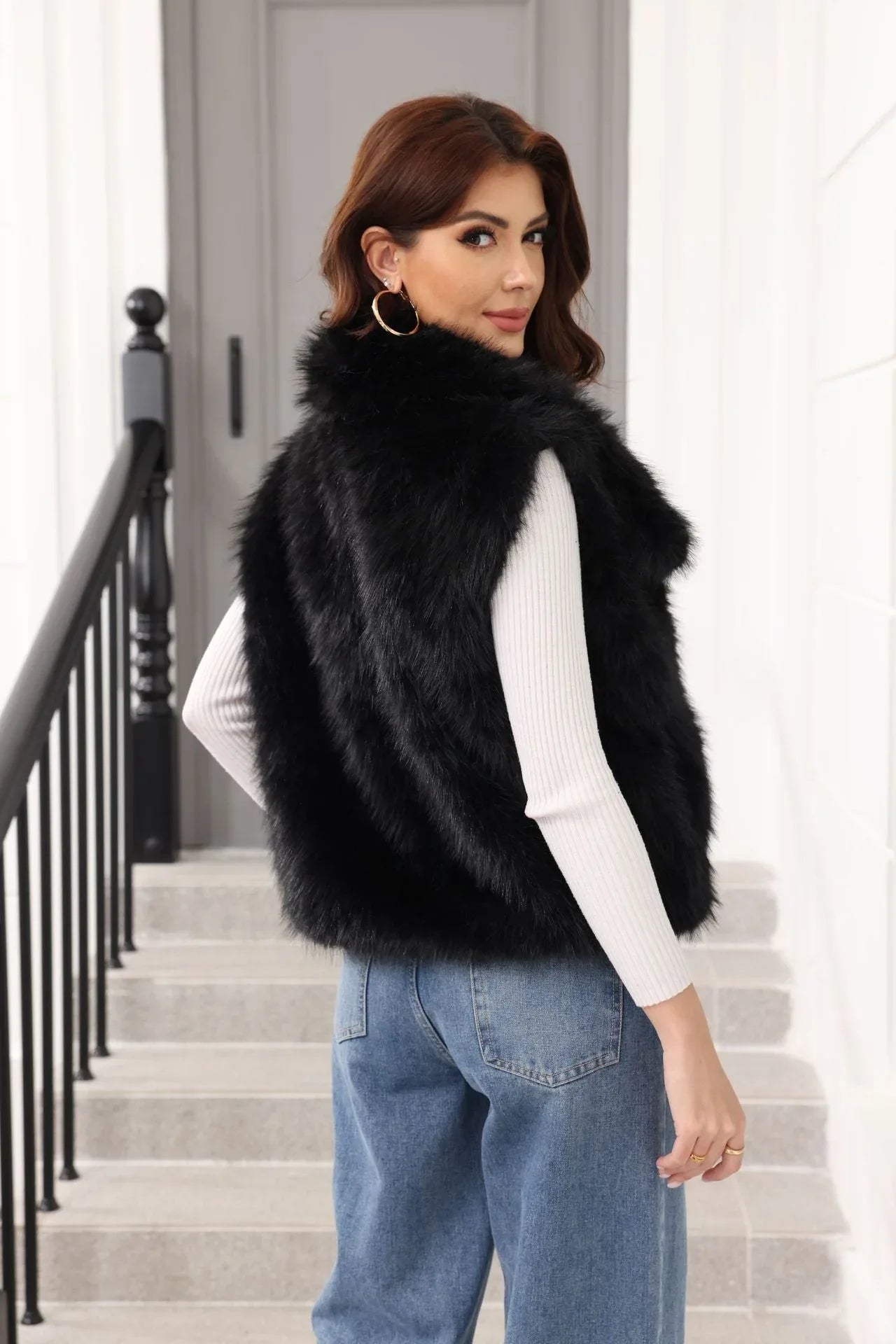 Woman wearing a black faux fur vest over a white top and blue jeans, turning to look over her shoulder.