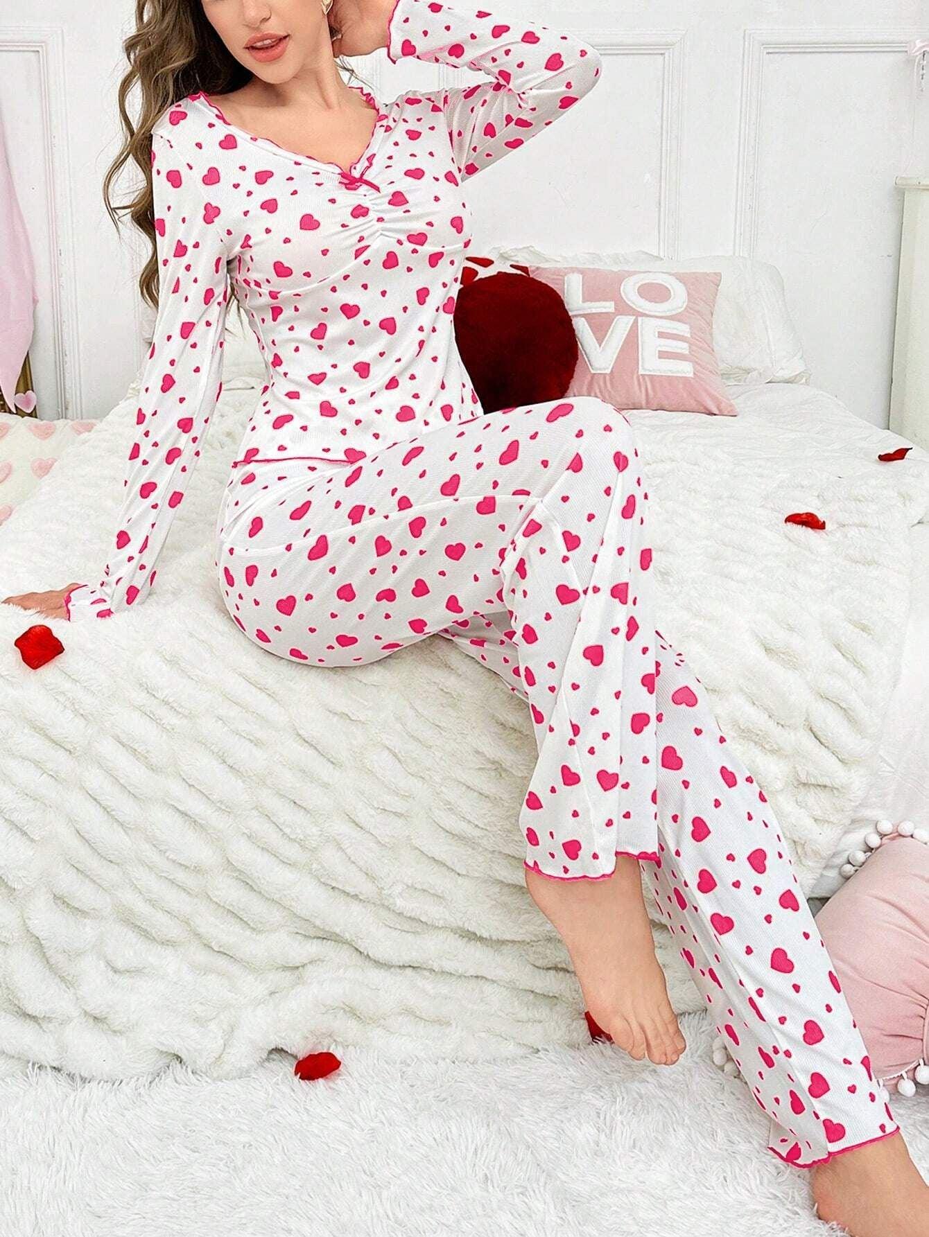 Woman sitting on a bed wearing a white long-sleeved pajama top and pants with a pink heart print, resting her hand on her chin.