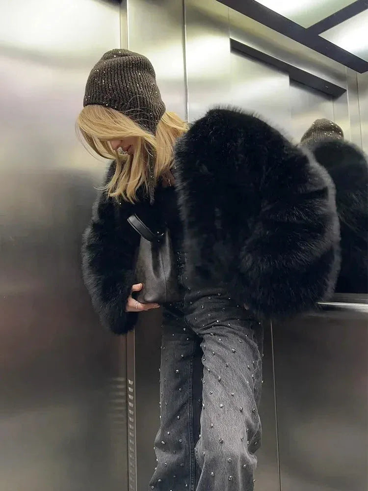 Woman wearing a black faux fur bomber jacket and a beanie, holding a handbag in an elevator.