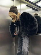 Woman wearing a black faux fur bomber jacket and a beanie, holding a handbag in an elevator.