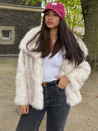 Woman in a cream faux fur jacket, dark jeans, and a pink baseball cap, posing outdoors in a park setting.