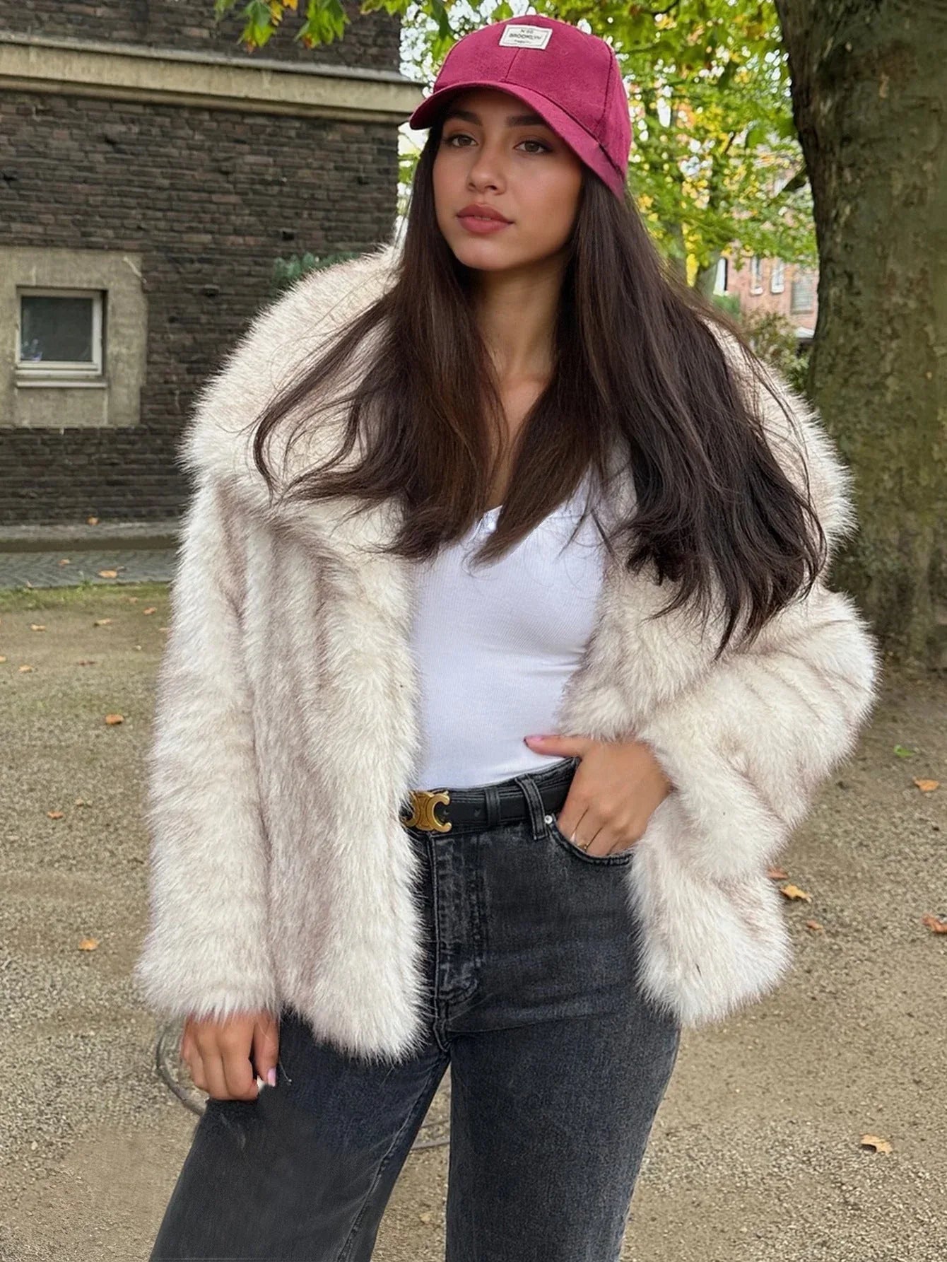 Woman in a cream faux fur jacket, dark jeans, and a pink baseball cap, posing outdoors in a park setting.