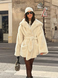 Woman walking in a street wearing a cream-colored faux fur coat with a hood, belt, and beanie.