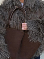 Close-up of a woman's hands holding a large brown tote bag, with the textured brown faux fur of her jacket visible.