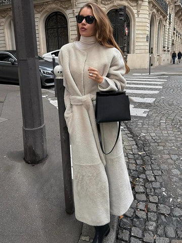 Woman wearing a long, cream-colored sherpa fleece coat with a belted waist and stand collar, standing on a city street.