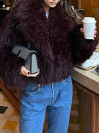 Woman holding a coffee and a bedazzled handbag, wearing a burgundy faux fur jacket and blue denim jeans.