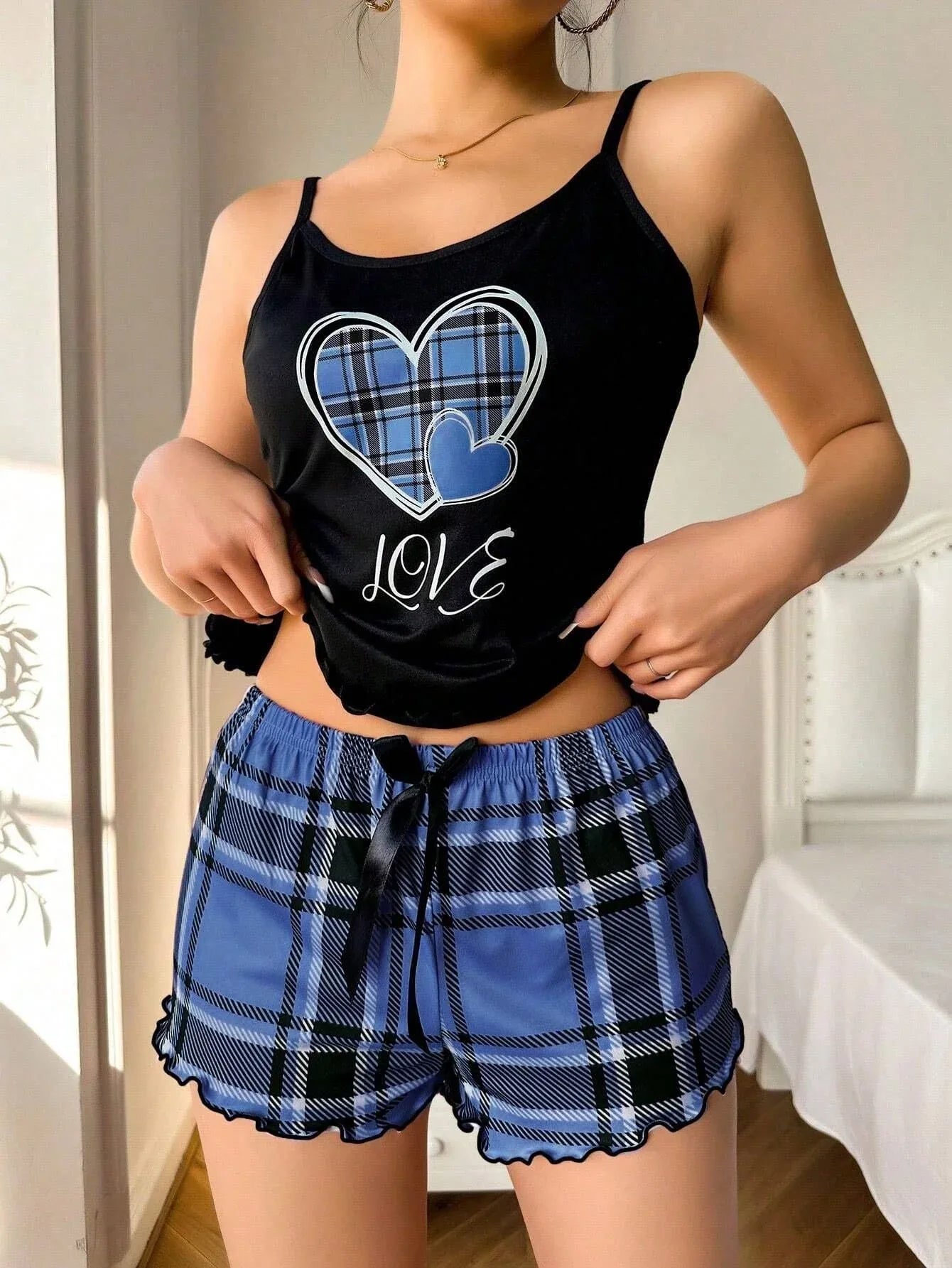 Woman wearing a black sleeveless pajama top with blue plaid heart graphic and 'love' text, and blue plaid pajama shorts with ruffled hem, standing and adjusting the pajama top.