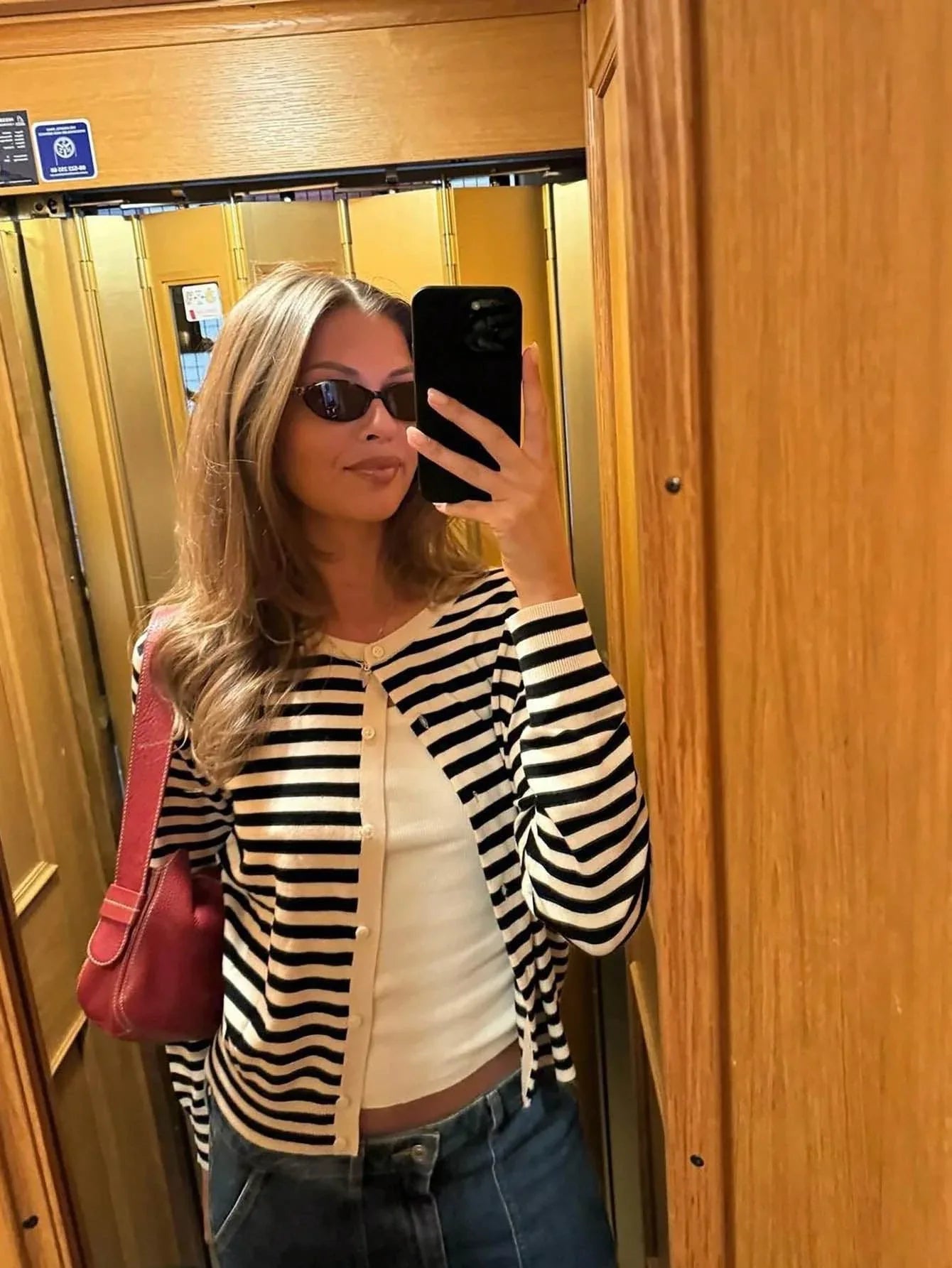 Woman taking a mirror selfie in an elevator, wearing a black and white striped cardigan, white tank top, and blue jeans.
