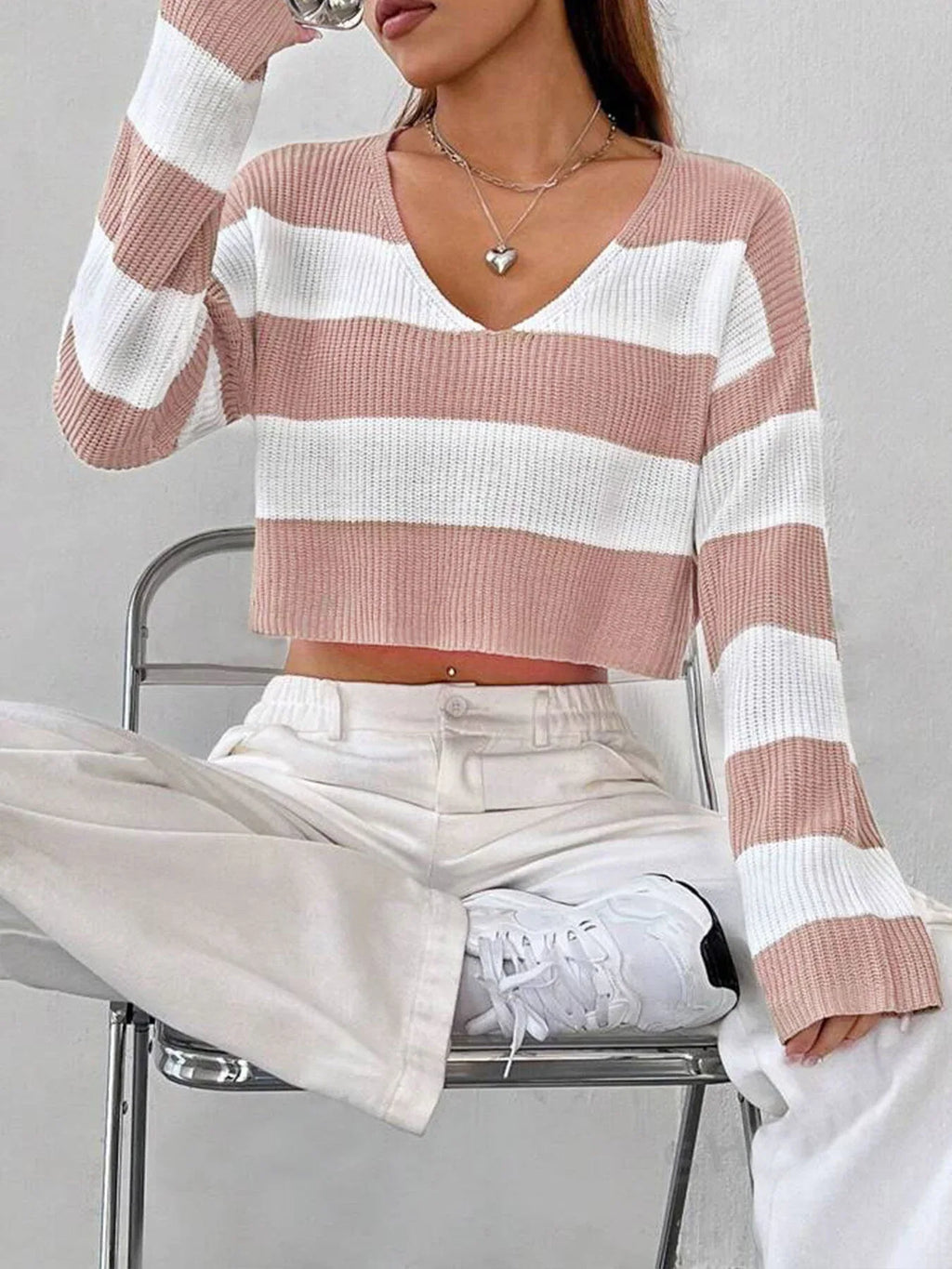 Woman sitting on a chair wearing a pink and white striped cropped knit sweater with white pants.