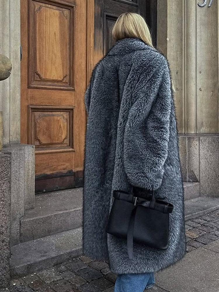 Rear view of a woman in a long grey faux fur coat with a lapel collar, walking outdoors.
