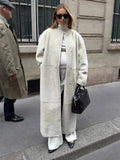 Woman in a cream sherpa coat and white trousers, accessorized with a black handbag and pointed-toe shoes.