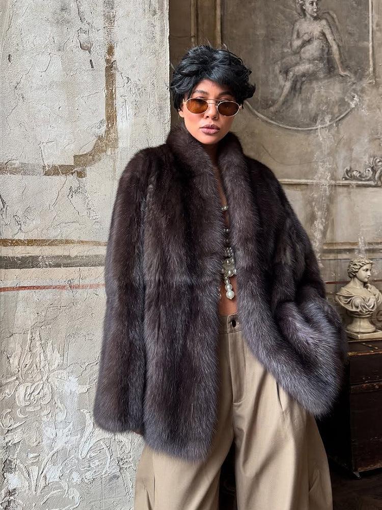 Model posing in a dark brown fur coat, tan trousers, and pearl necklace.