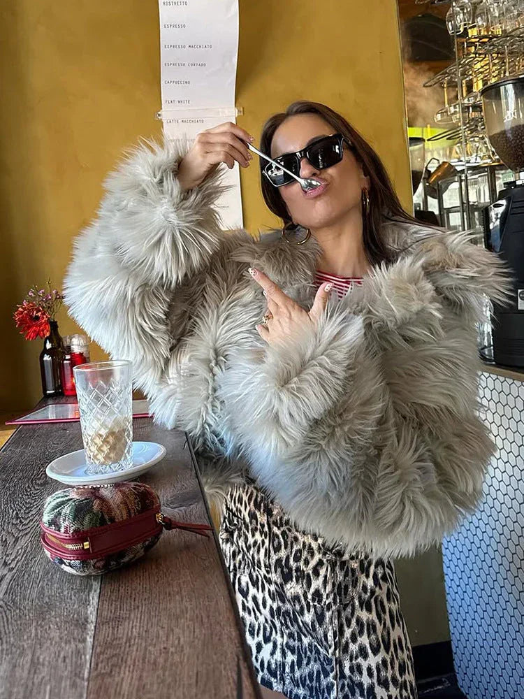 Woman in a light grey faux fur jacket posing at a cafe table