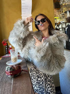 Woman in a light grey faux fur jacket posing at a cafe table