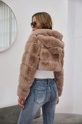 Back view of a woman in a cropped taupe faux fur hooded jacket and blue jeans.