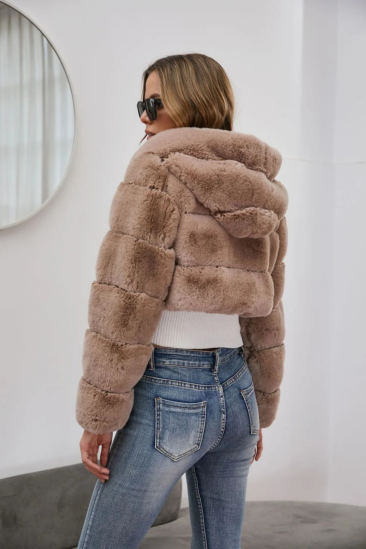 Back view of a woman in a cropped taupe faux fur hooded jacket and blue jeans.