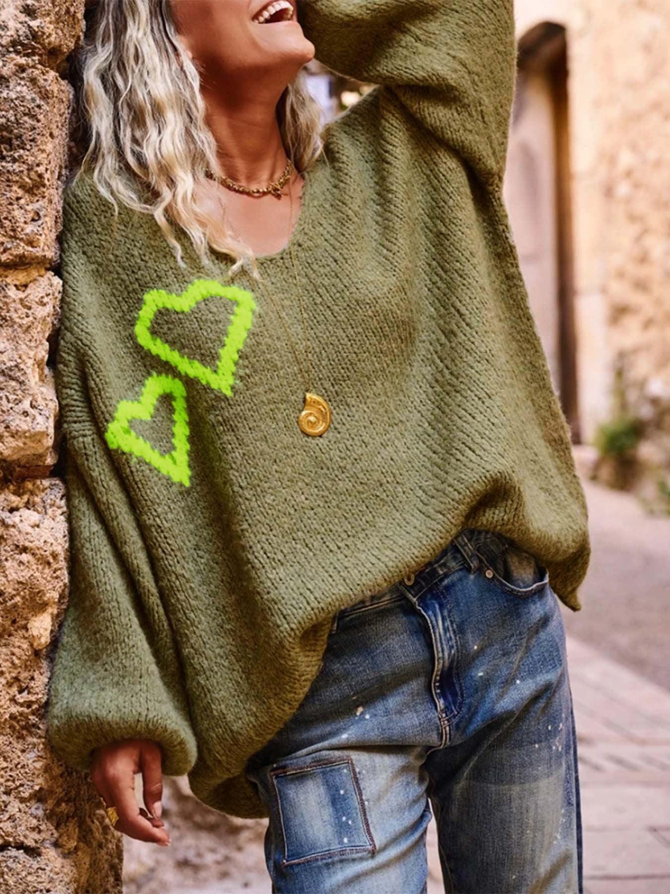 Three-quarter view of a woman smiling and wearing the olive green V-neck knit sweater with heart embroidery.