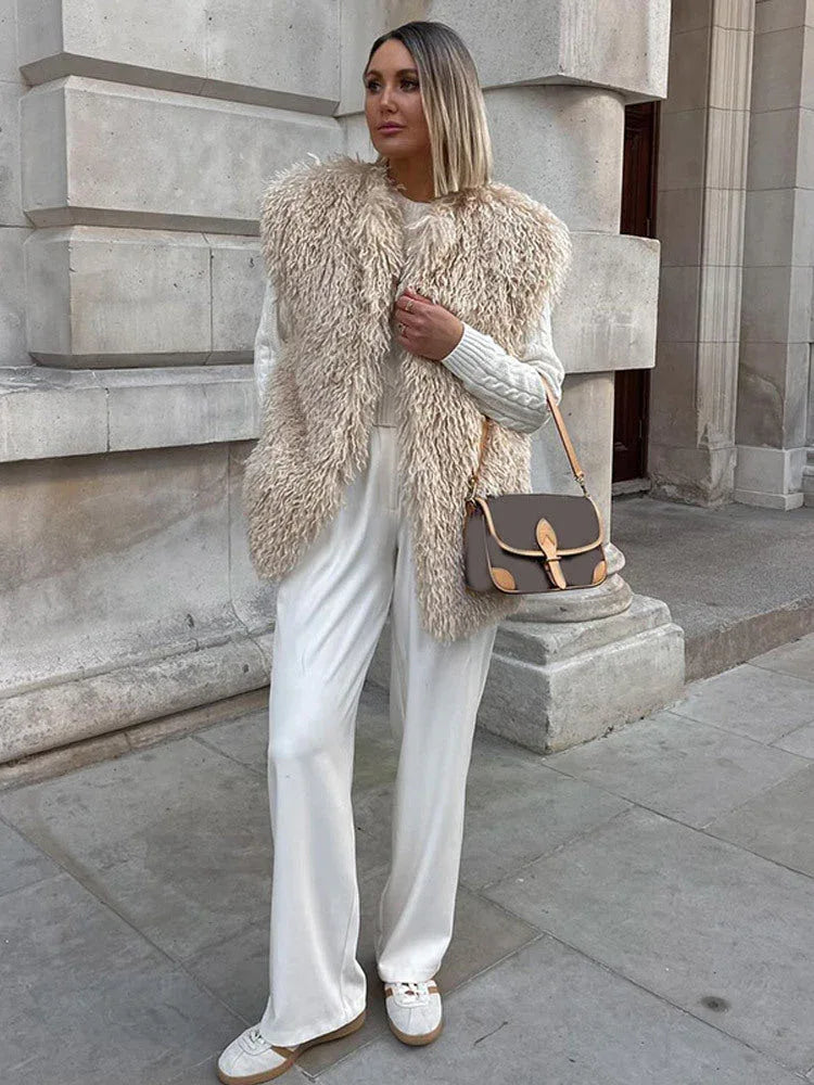 Woman wearing a beige faux fur vest over a white cable-knit sweater and white wide-leg pants with sneakers