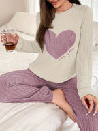 Woman sitting on a bed, wearing a cream long-sleeve pajama top with a pink heart graphic and lilac ribbed pajama pants, holding a drink.