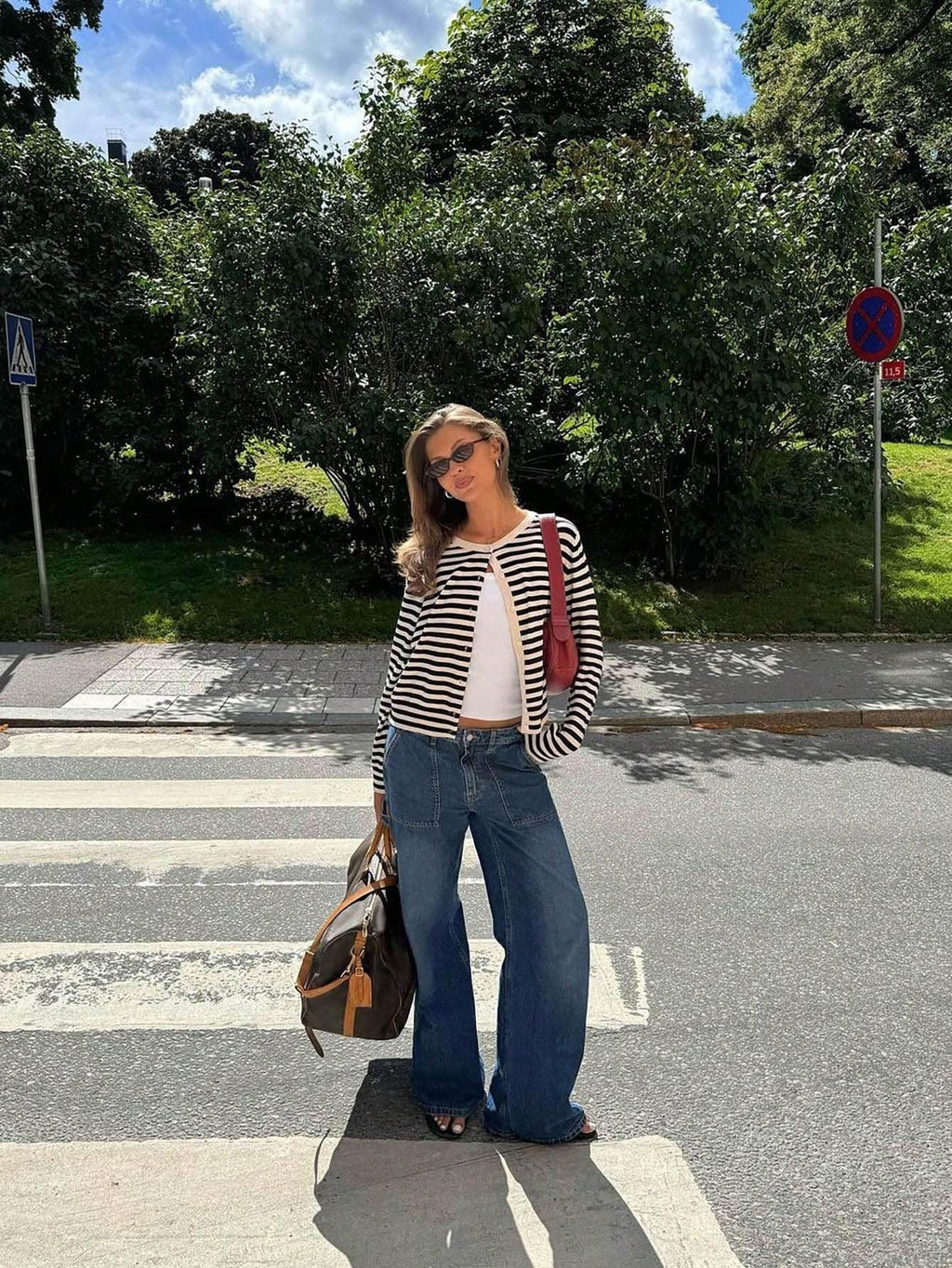 Woman in a black and white striped cardigan, white tank top, and blue wide-leg jeans holding a duffel bag on a crosswalk.