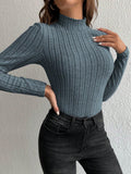 Woman touching her chest while wearing a grey ribbed long-sleeve mock neck top and black jeans