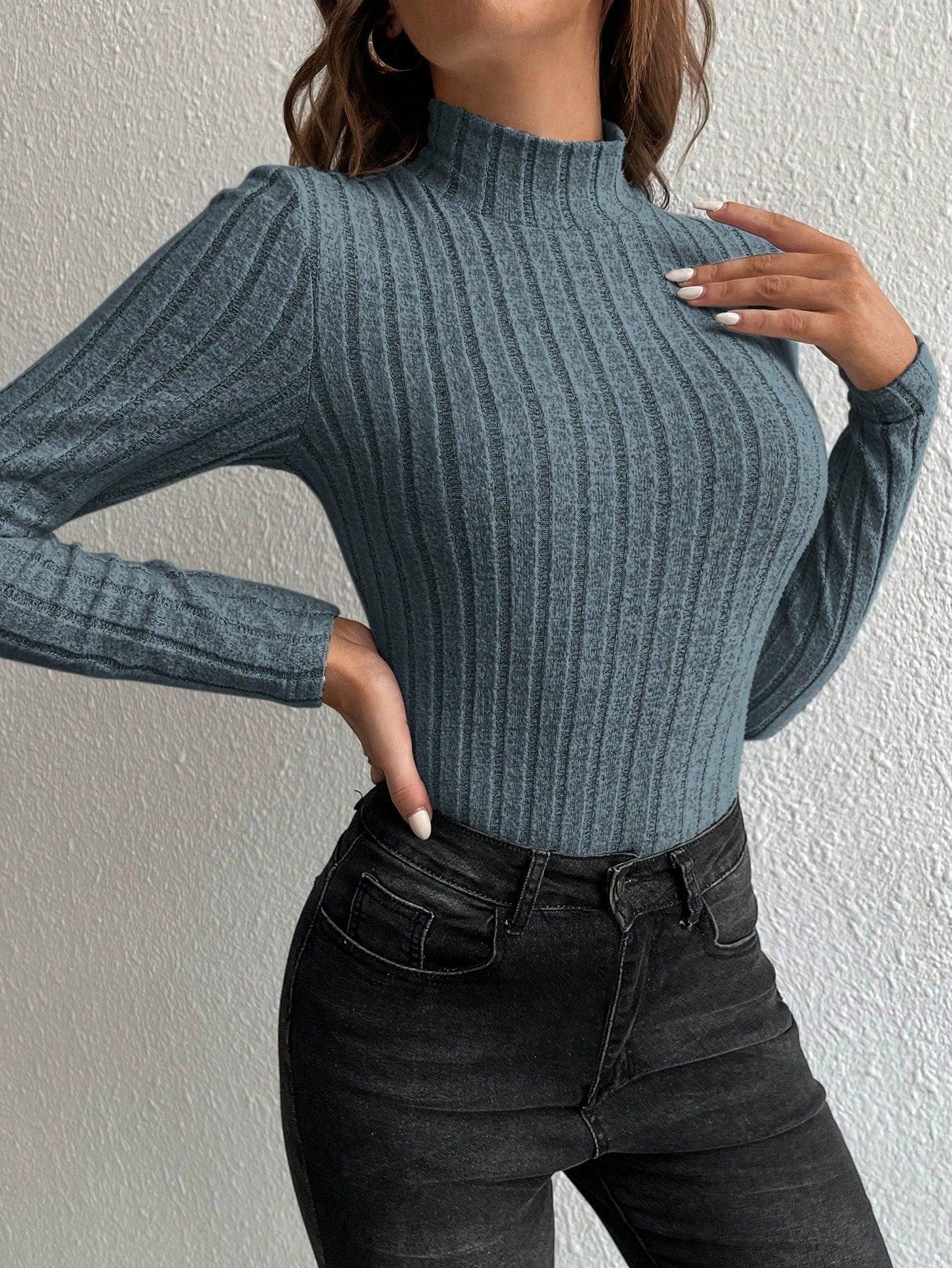 Woman touching her chest while wearing a grey ribbed long-sleeve mock neck top and black jeans