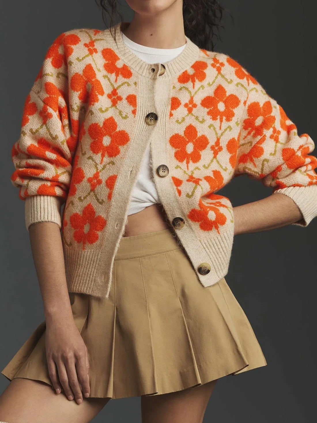Woman wearing a beige cardigan with an orange floral print, a white t-shirt, and a tan pleated mini skirt.