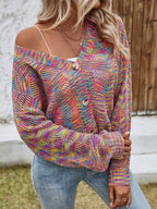 Close-up of a woman wearing a rainbow multicolor open-knit cropped cardigan with a v-neck and button-front closure, layered over a white top and blue jeans, with one shoulder strap visible.