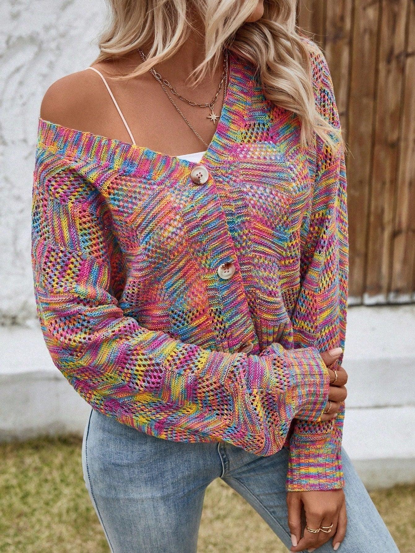 Close-up of a woman wearing a rainbow multicolor open-knit cropped cardigan with a v-neck and button-front closure, layered over a white top and blue jeans, with one shoulder strap visible.