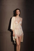 Woman wearing a cream satin camisole and shorts set with pink lace trim and a matching cream satin robe, standing and looking forward.