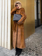 Woman posing in a long brown faux fur coat with a black quilted handbag.