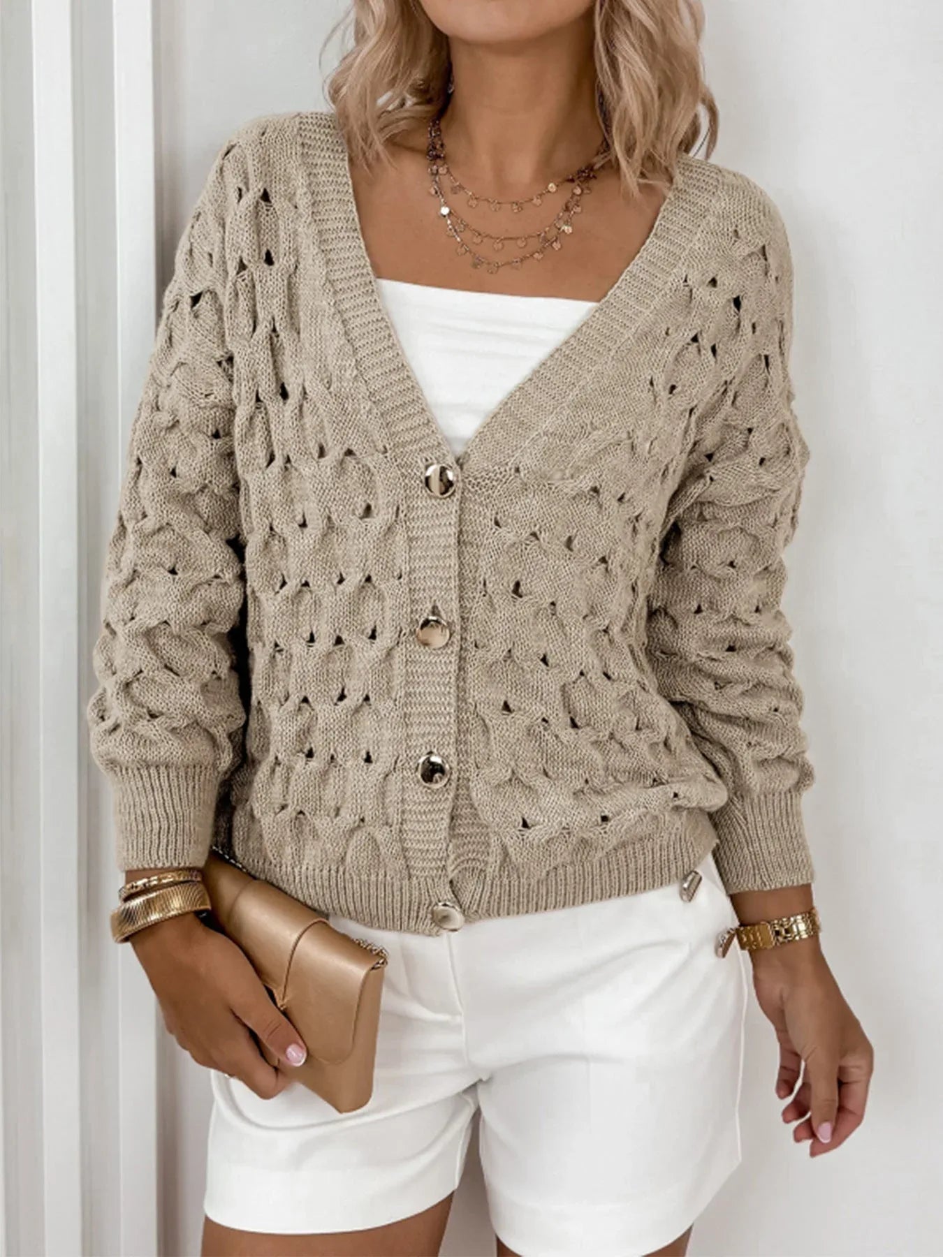 Model styling a beige open-knit cardigan with white shorts and gold accessories.